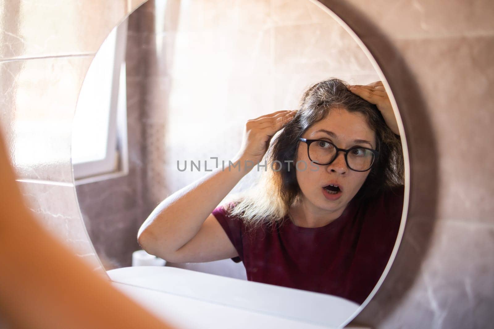 Gray haired surprised caucasian middle aged woman looking at grey hair head in mirror reflection. Early gray hair concept by Satura86