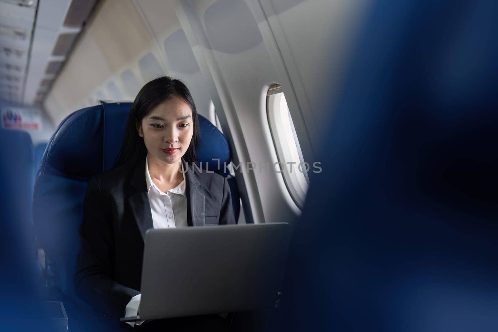 A woman is sitting in an airplane seat with a laptop open in front of her. She is wearing a suit and she is working on her laptop. Concept of productivity and focus