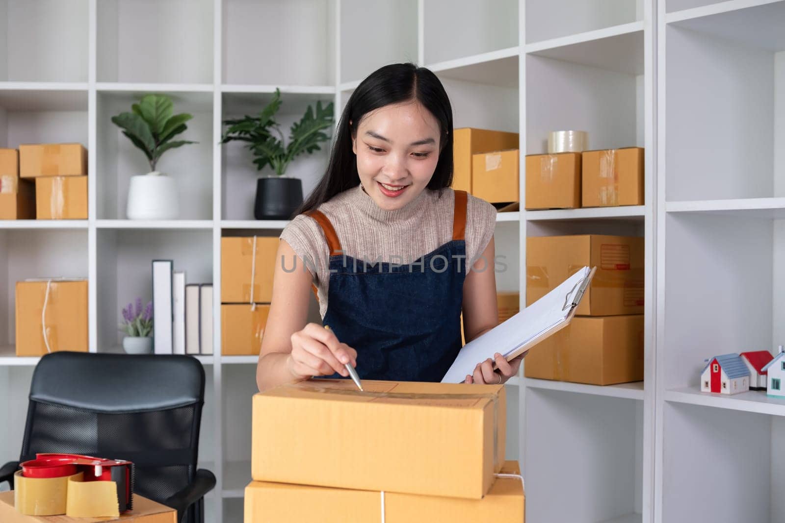 A small business owner receives product orders and writes shipping information on cardboard boxes in the home office to prepare for delivery. by wichayada