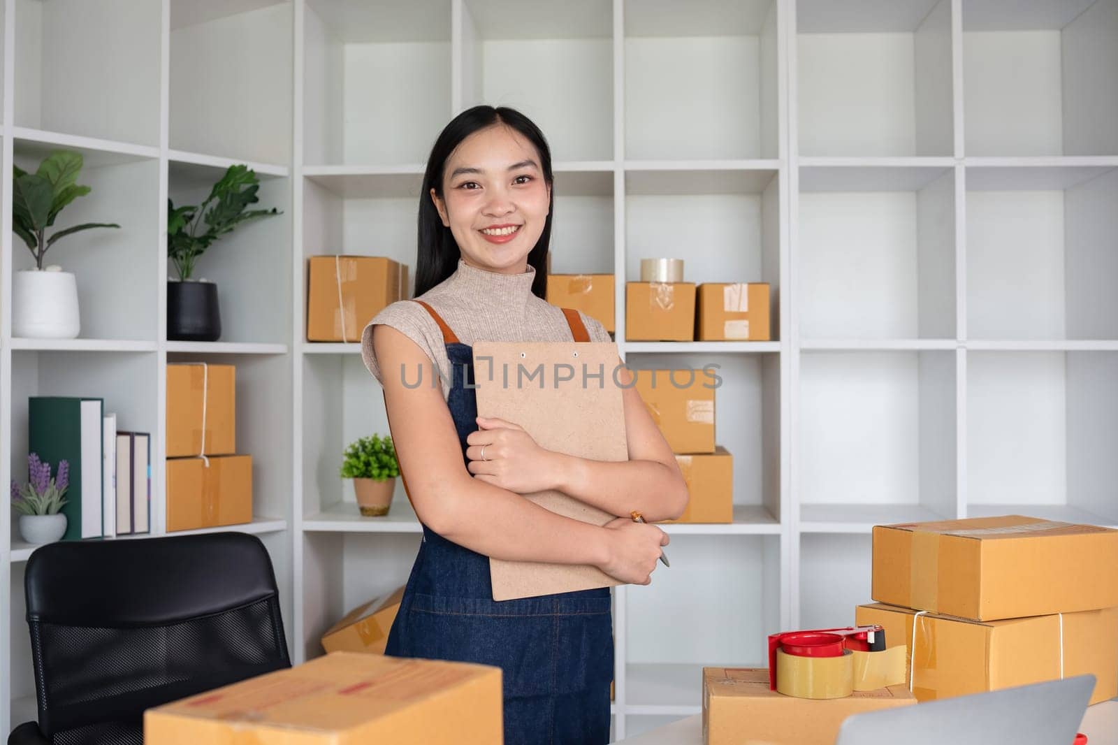 Confident owner of a small online sales business stands holding a clipboard with inventory and shipping information in his home office. Small entrepreneurs prepare to deliver to customers. by wichayada