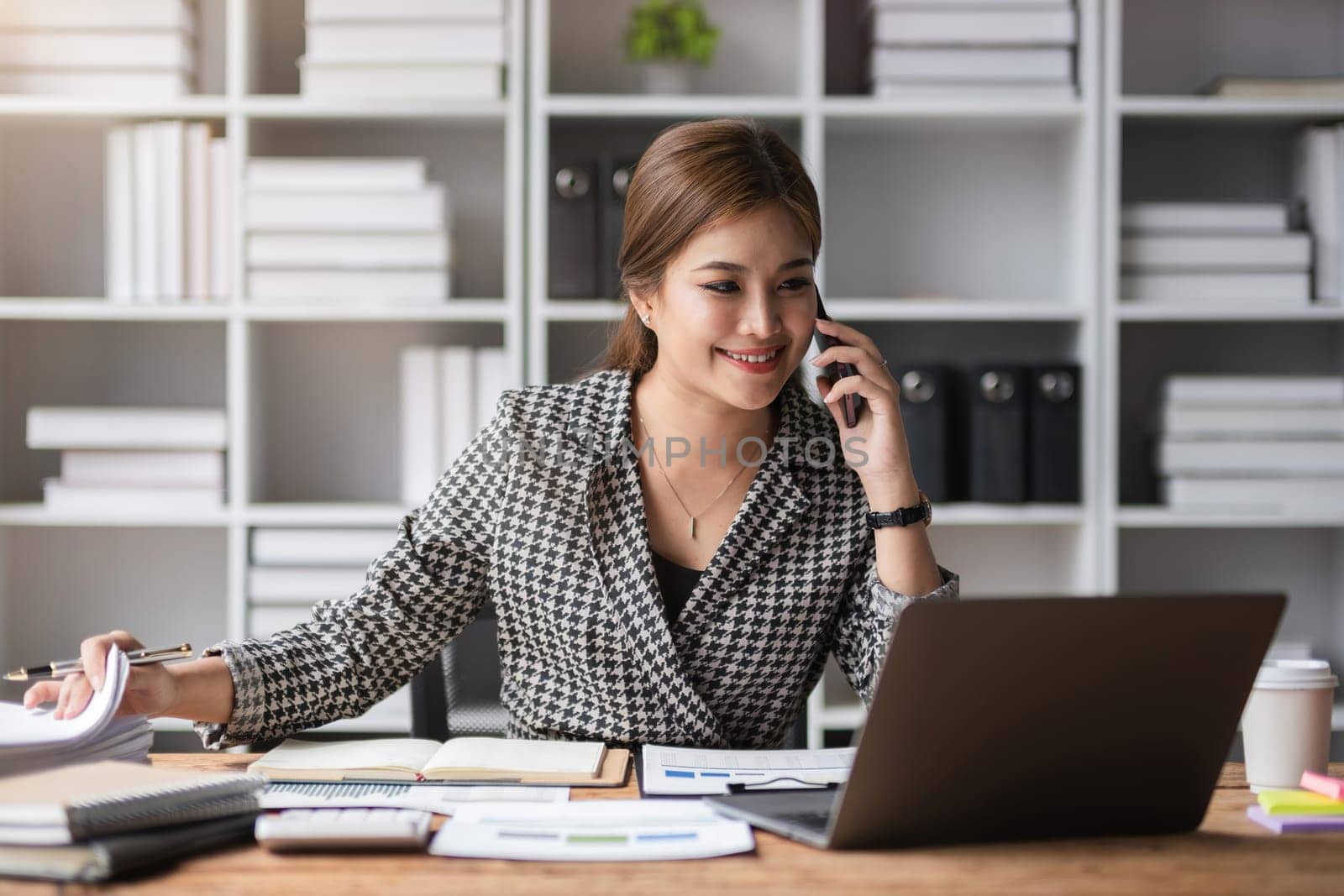 Asian female accountant is talking on the phone in the office with working documents regarding calculations on the office table. by wichayada