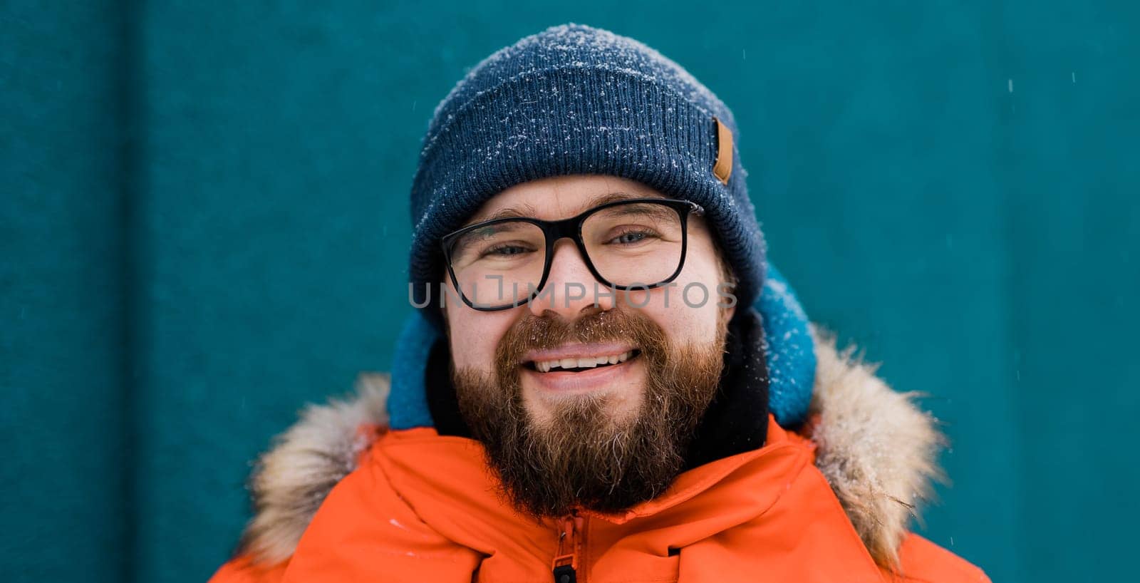 Banner portrait handsome eyeglasses young male smiling bearded stands on a blue wall background in a bright orange winter jacket with a hood with fur in winter. Millennial generation and Gen Y concept by Satura86