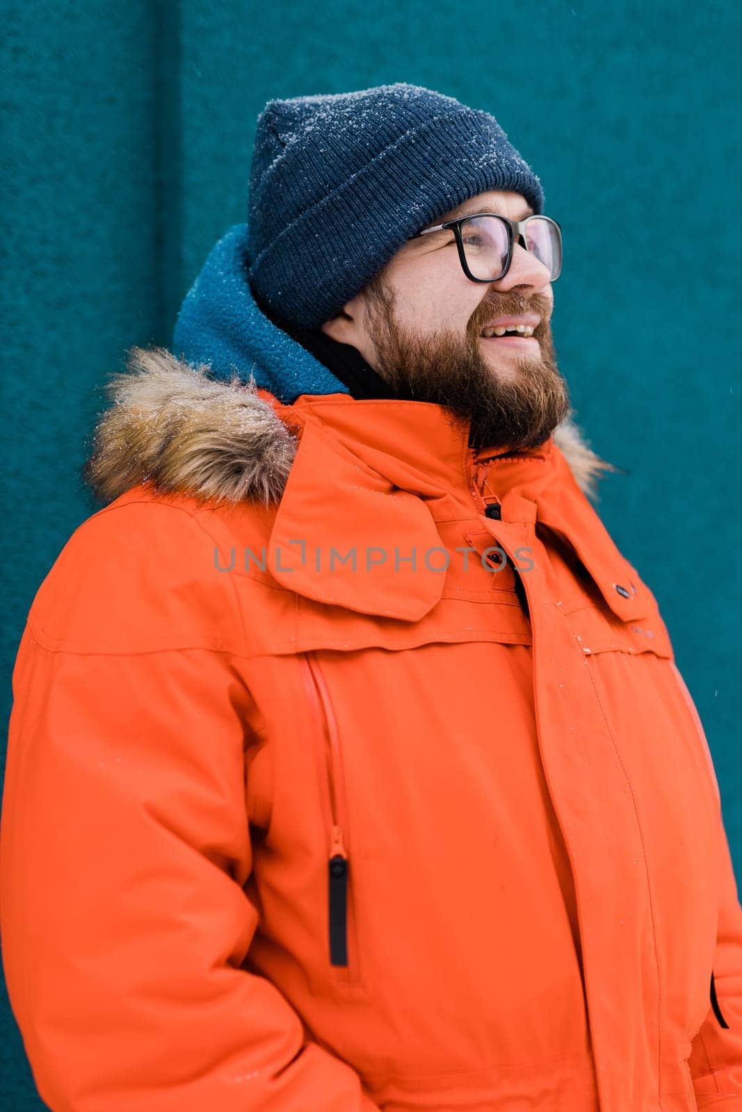 Portrait handsome eyeglasses young male smiling bearded stands on a blue wall background in a bright orange winter jacket with a hood with fur in winter. Millennial generation and Gen Y