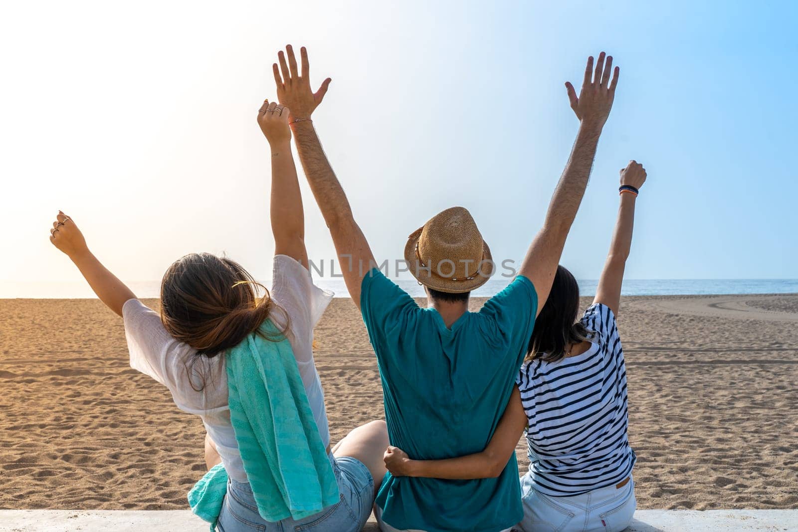 Happy friends holding each other in the beach with raised arms. Cheerful embracing each other, back view. High quality photo