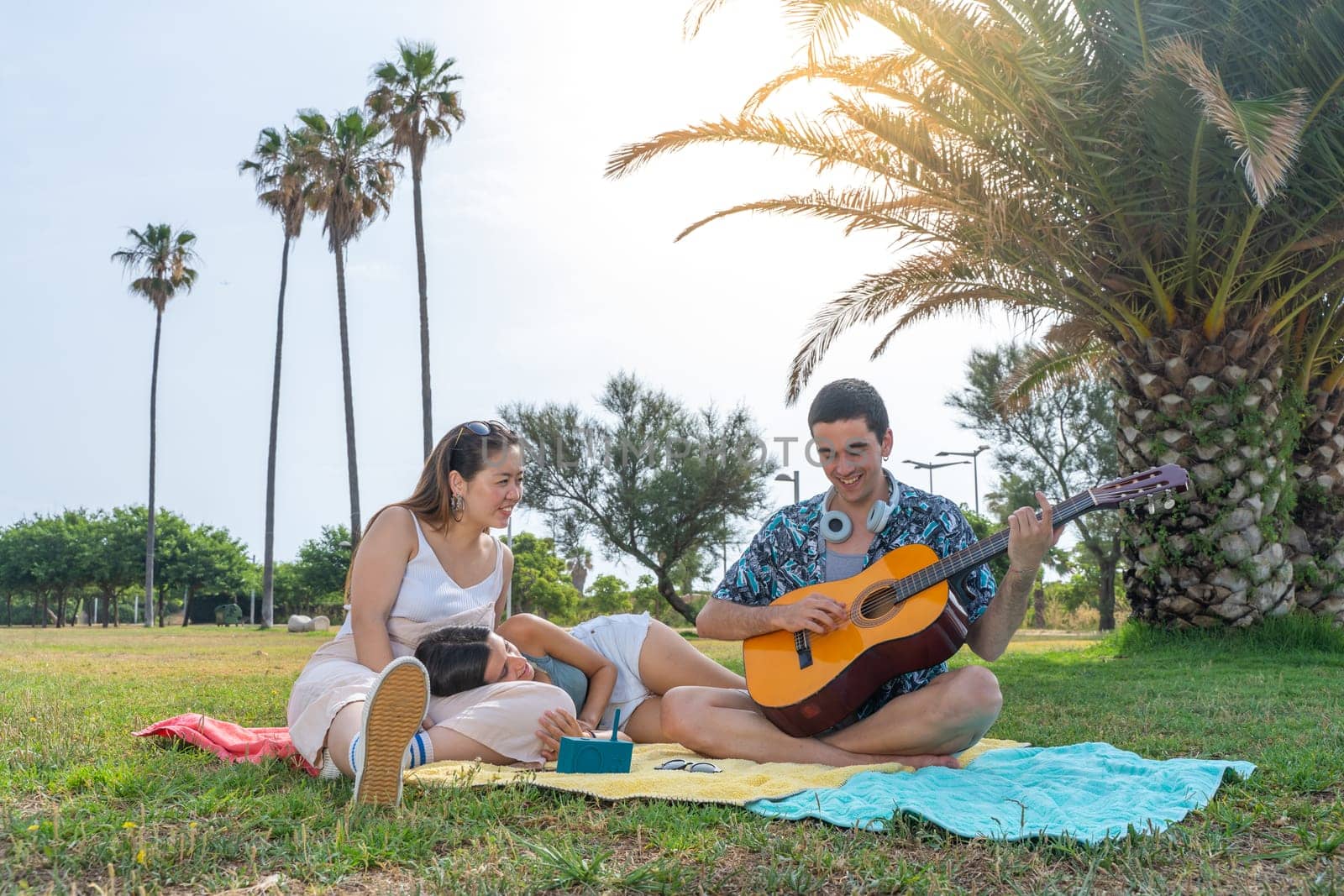 Multiracial teenagers playing guitar while sitting on green lawn in park and enjoying summer day together. High quality photo