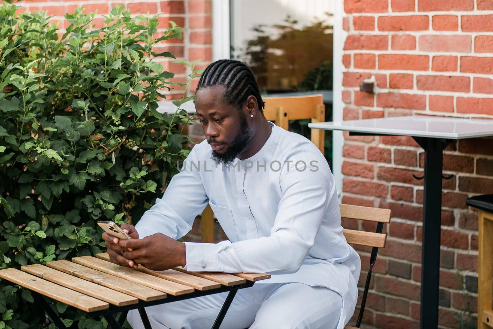 African american man in stylish ethnic dashiki wear viewing photos in social networks on mobile enjoying free time outdoors. Dark-skinned man watching funny video on smartphone and social networks. Blog and blogger scrolling content in Internet by Satura86