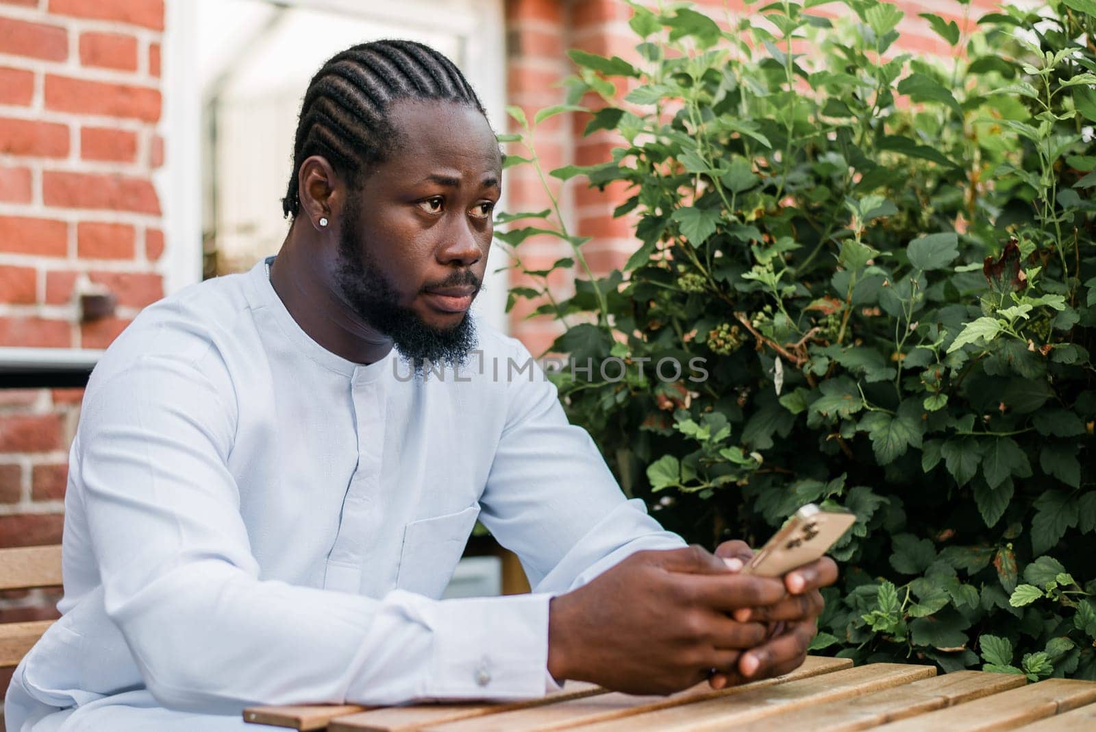 African American hipster guy in cultural and ethnic clothing dashiki chatting on smartphone outdoors, texting with friends on social networks. Millennial generation and Internet blog concept by Satura86