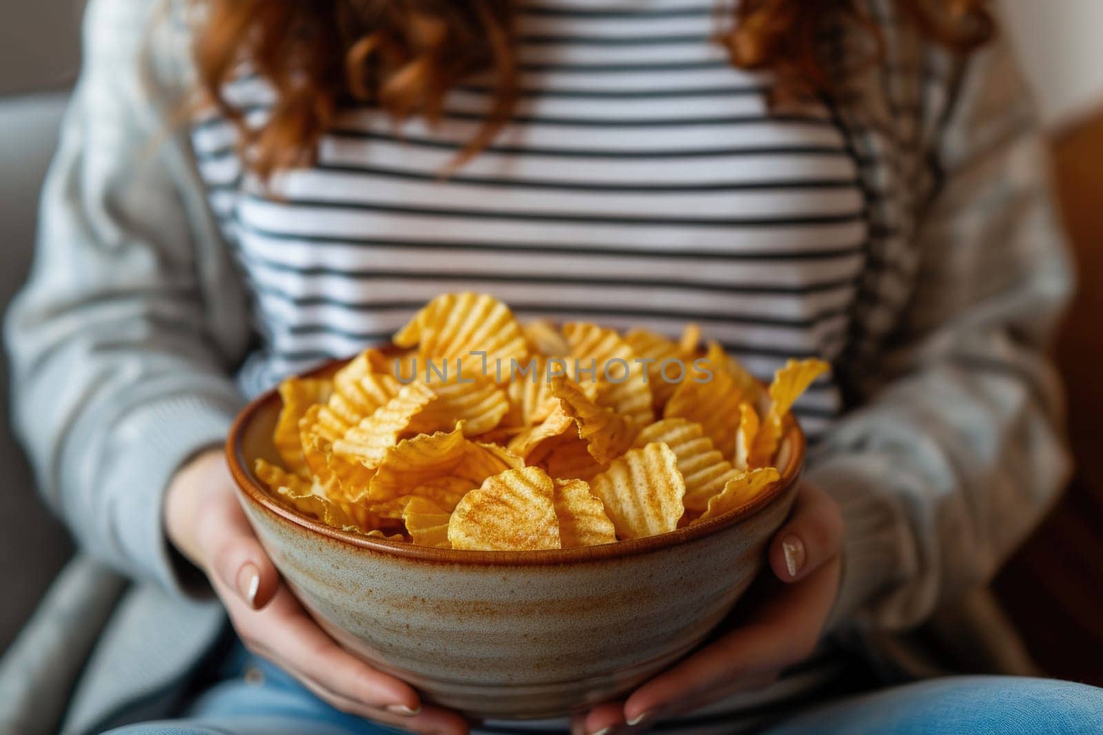 Pregnant woman sitting on the sofa enjoys eating potato chips from a bowl at home. ai generated by Desperada