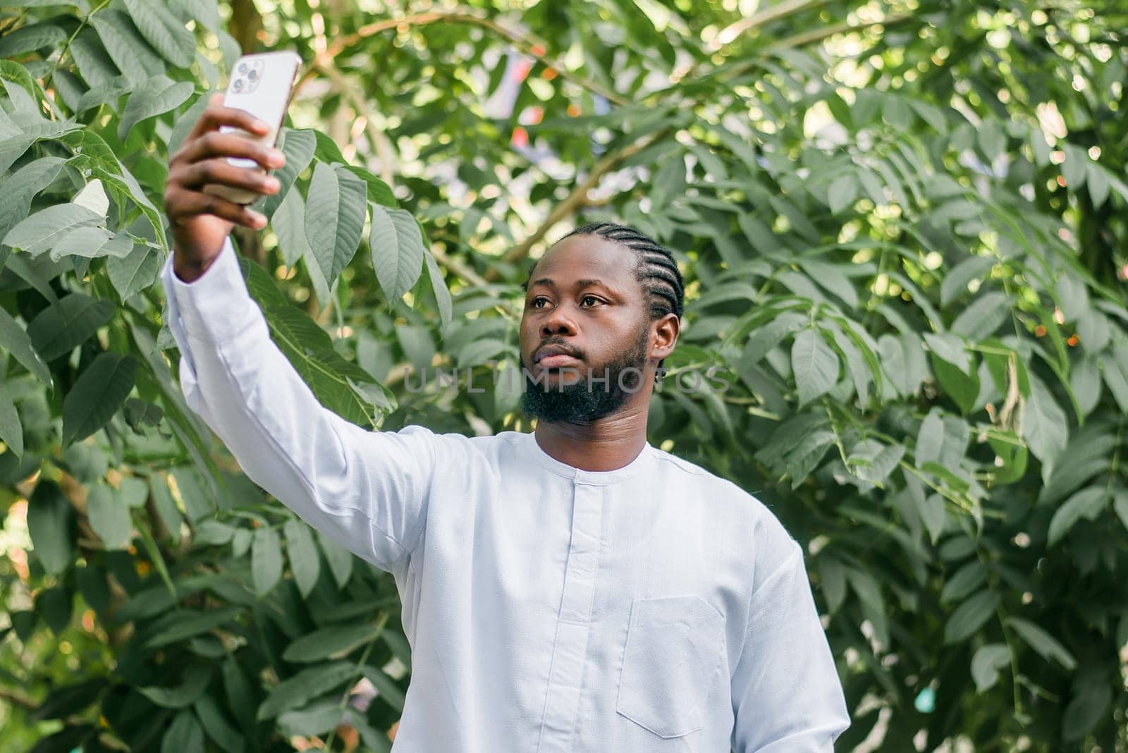 Happy young African American man in dashiki ethnic clothes taking selfie on green leaves outdoor background. Millennial generation student and youth. by Satura86