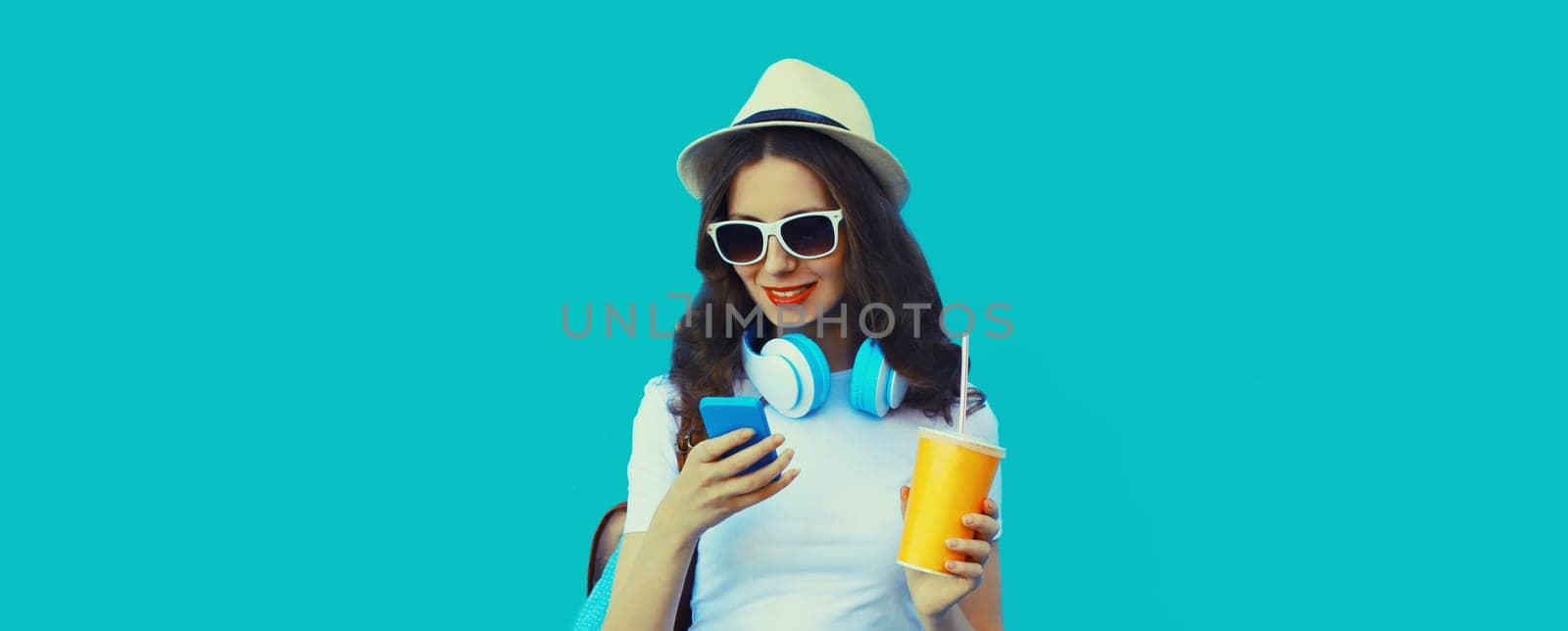 Portrait of modern young woman listening to music with headphones holding phone and cup of fresh juice on blue studio background