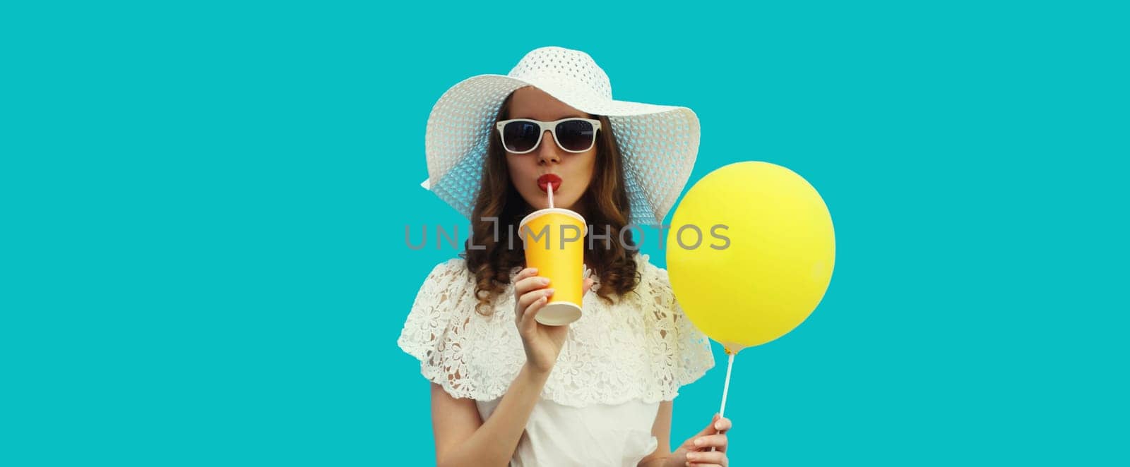 Portrait of beautiful young woman model with cup of coffee or fresh juice holding yellow balloon wearing white summer straw hat on studio blue background