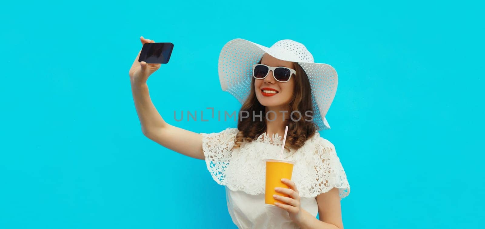 Summer portrait of beautiful happy smiling young woman taking selfie with mobile phone holding cup of juice wearing white straw hat on blue background