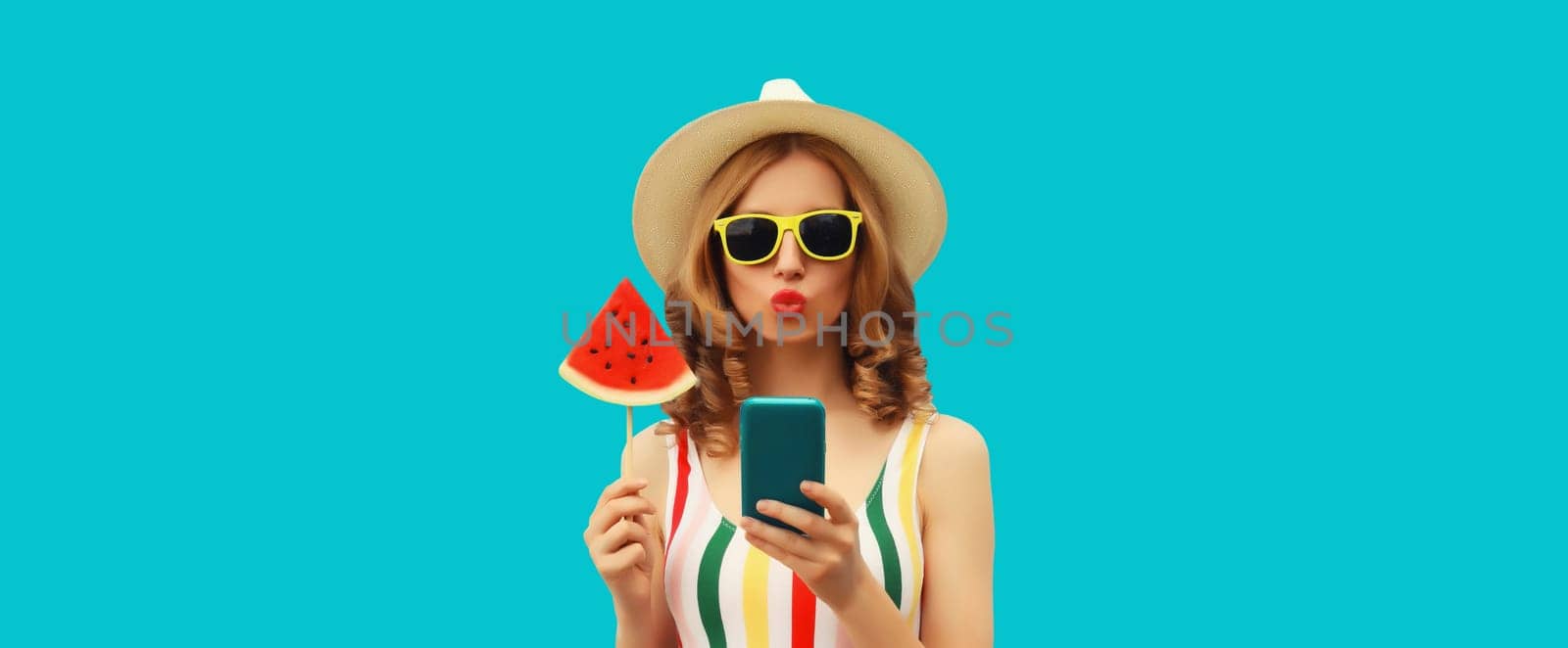 Summer portrait of happy young woman holding smartphone with juicy lollipop or ice cream shaped slice of watermelon wearing straw hat on blue background