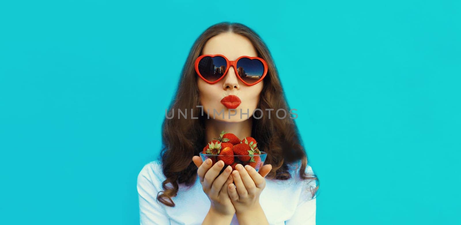 Portrait of beautiful caucasian young woman with handful of fresh strawberries blowing kiss wearing red heart shaped sunglasses on blue studio background