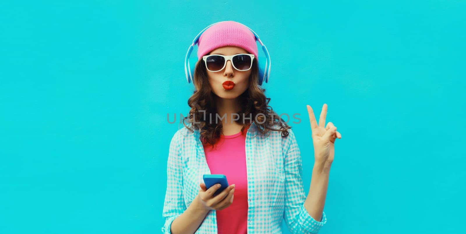 Portrait of stylish modern happy young woman listening to music in headphones with mobile phone wearing pink hat, sunglasses on blue studio background