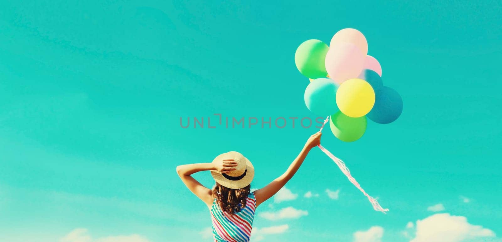 Back view of happy joyful young woman with bunch of colorful balloons in summer straw hat on blue sky clouds background