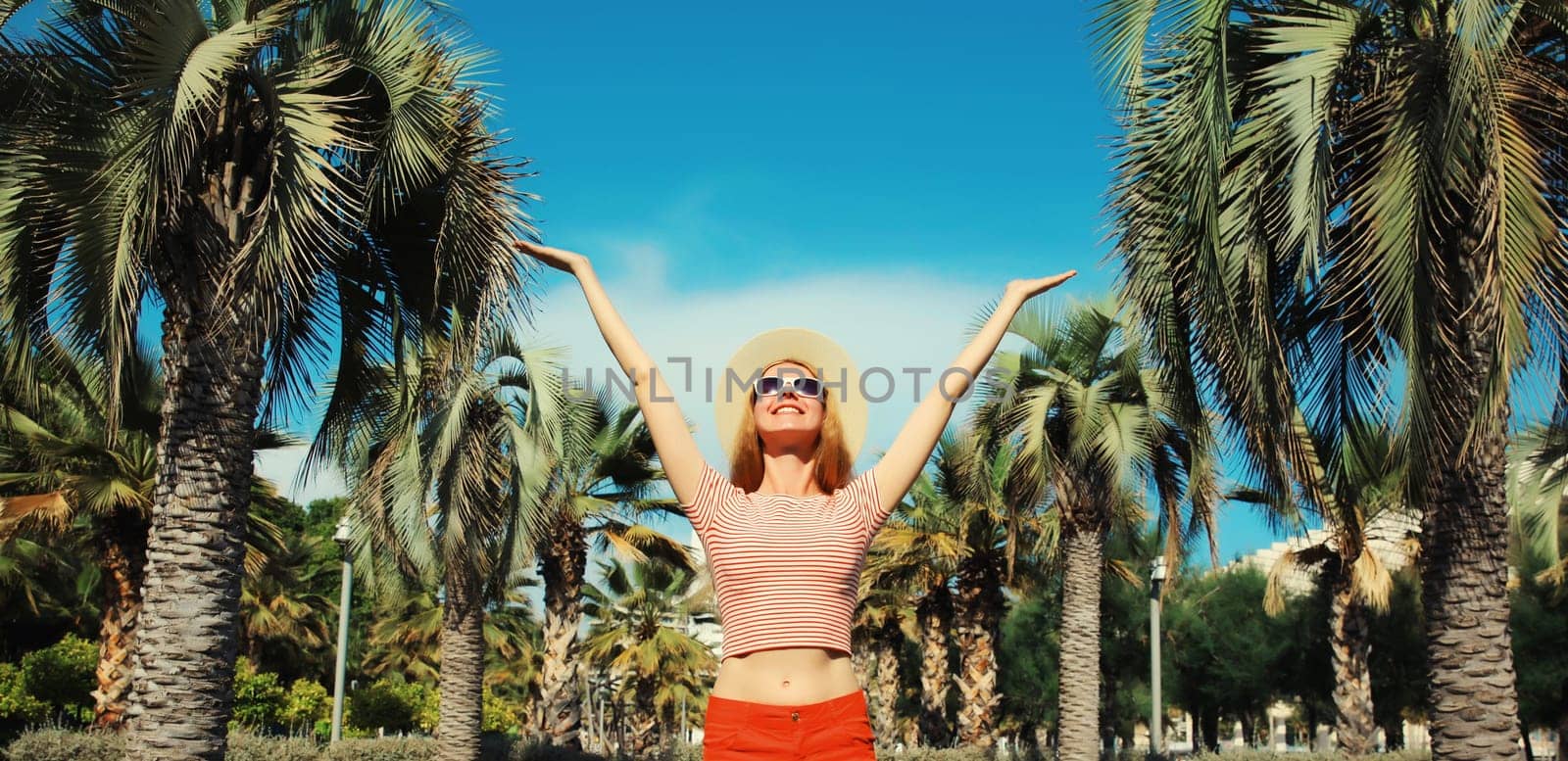 Summer vacation, happy cheerful young woman raising her hands up against a palm tree background
