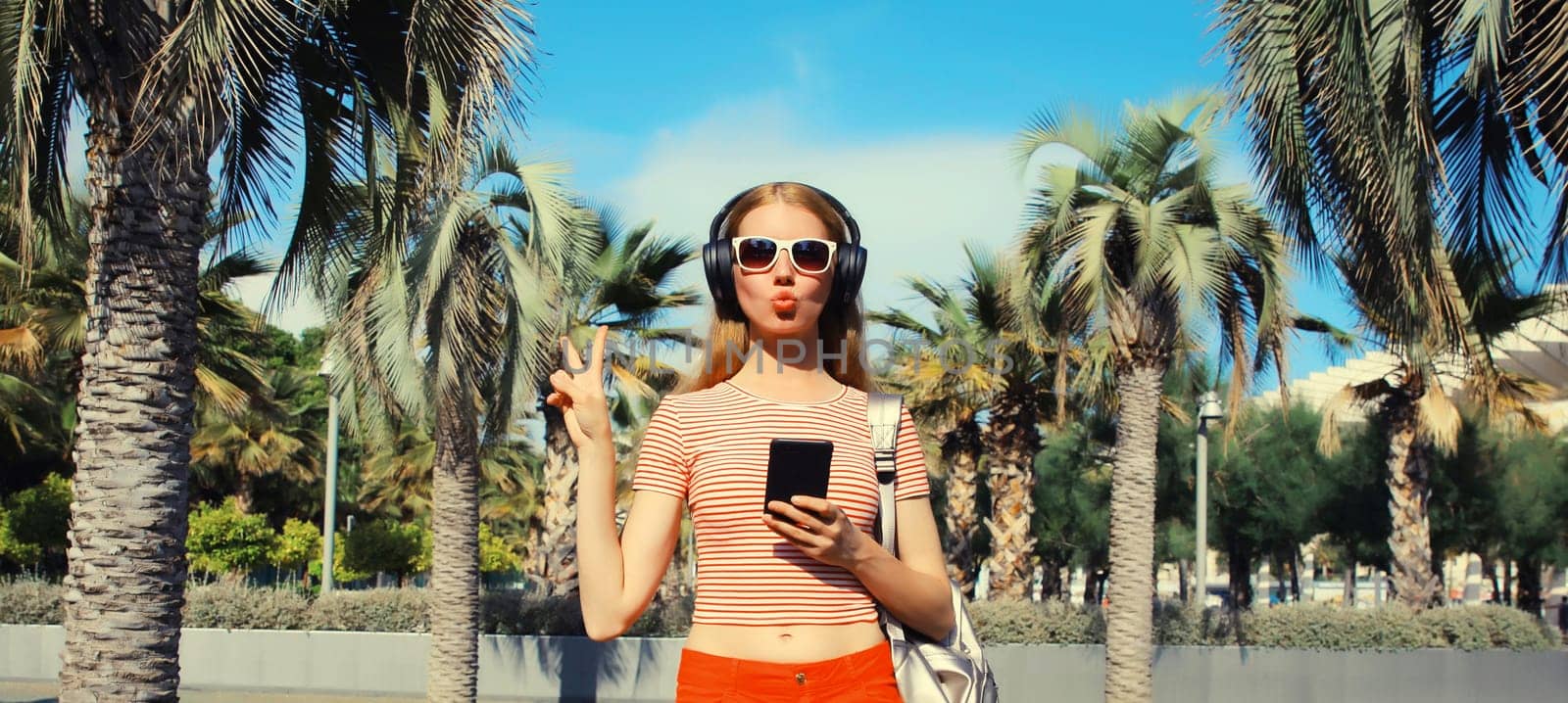 Happy smiling young woman listening to music in headphones with smartphone in summer park against a palm trees background