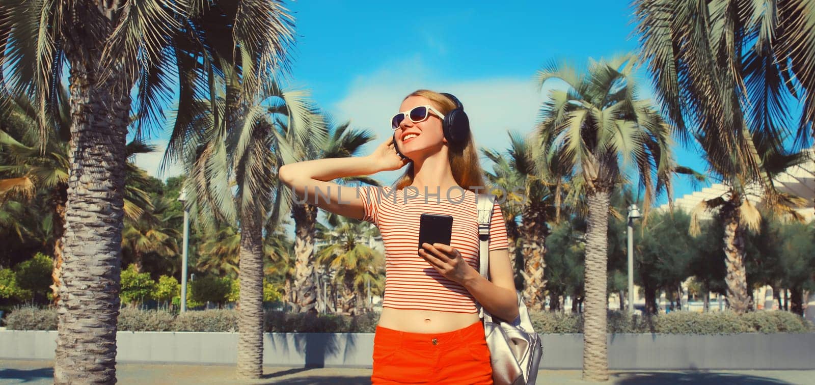 Happy smiling young woman listening to music in headphones with smartphone in summer park against a palm trees background
