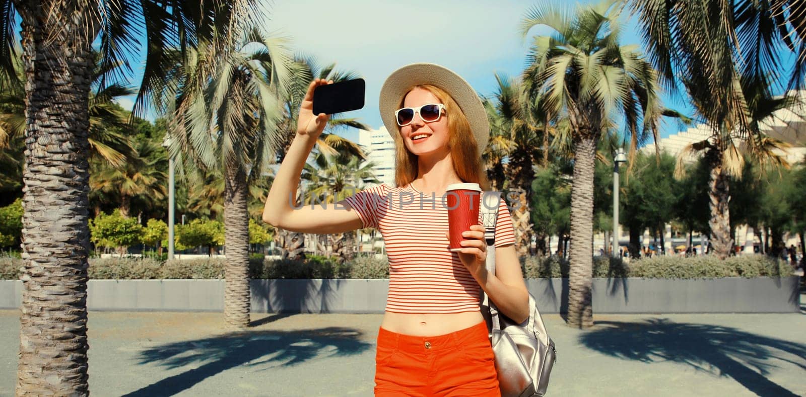 Portrait of young woman taking selfie with phone in summer park wearing backpack, straw hat against a palm trees background