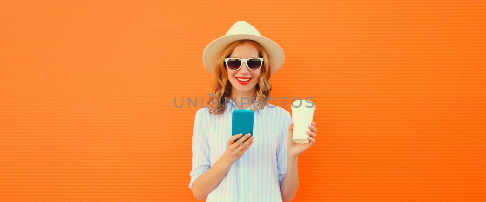 Summer portrait of happy traveler young woman with mobile phone looking at device in straw tourist hat on orange background