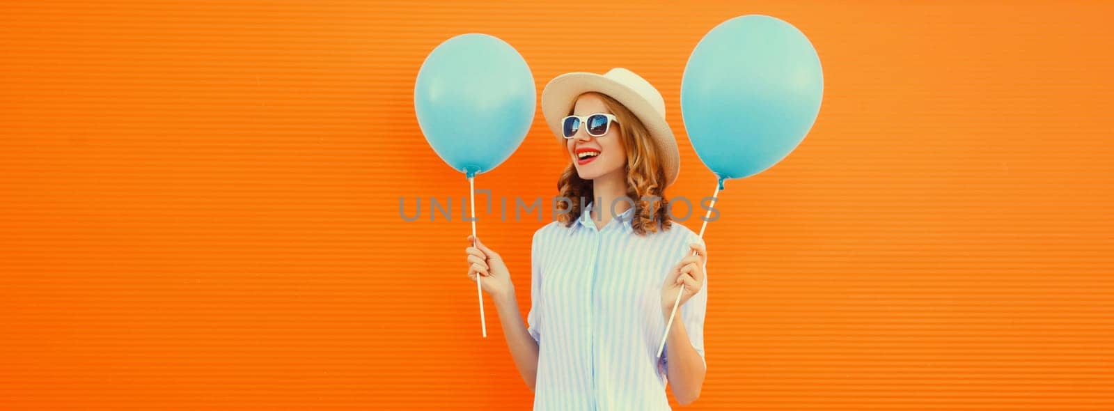 Portrait of happy smiling young woman with blue balloon wearing summer straw hat on orange background