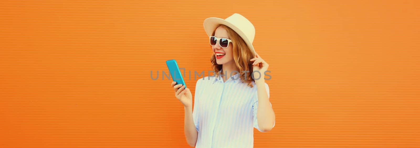 Summer portrait of happy traveler young woman with mobile phone looking at device in straw tourist hat on orange background