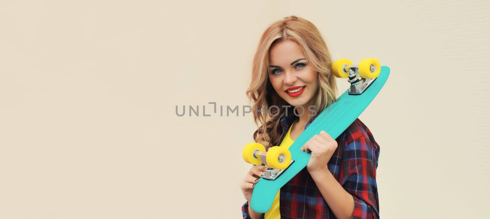 Portrait of stylish young blonde woman posing with skateboard on city street against white wall background, blank copy space