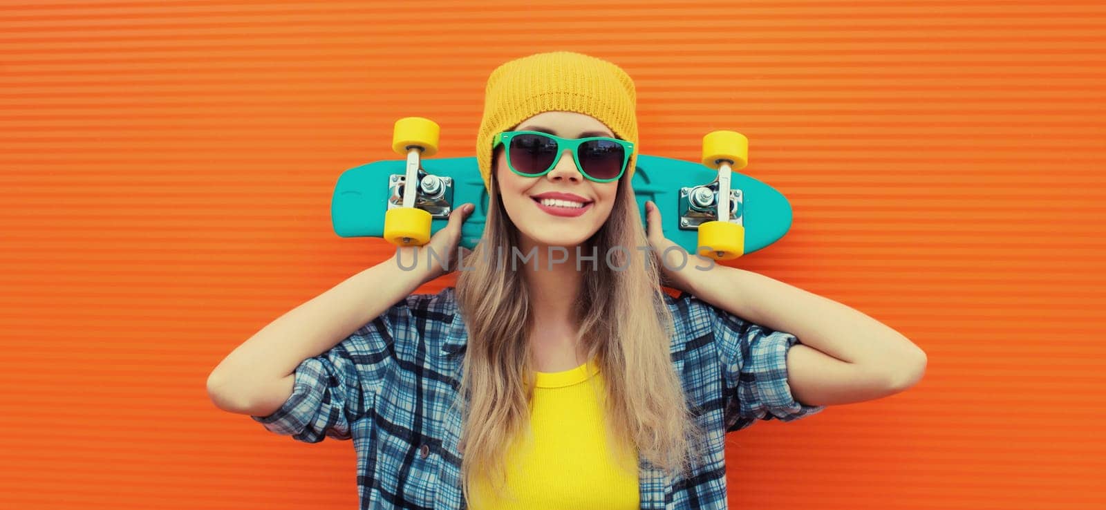 Summer portrait of happy cheerful stylish young woman with skateboard in colorful clothes, yellow hat posing on orange background on city street