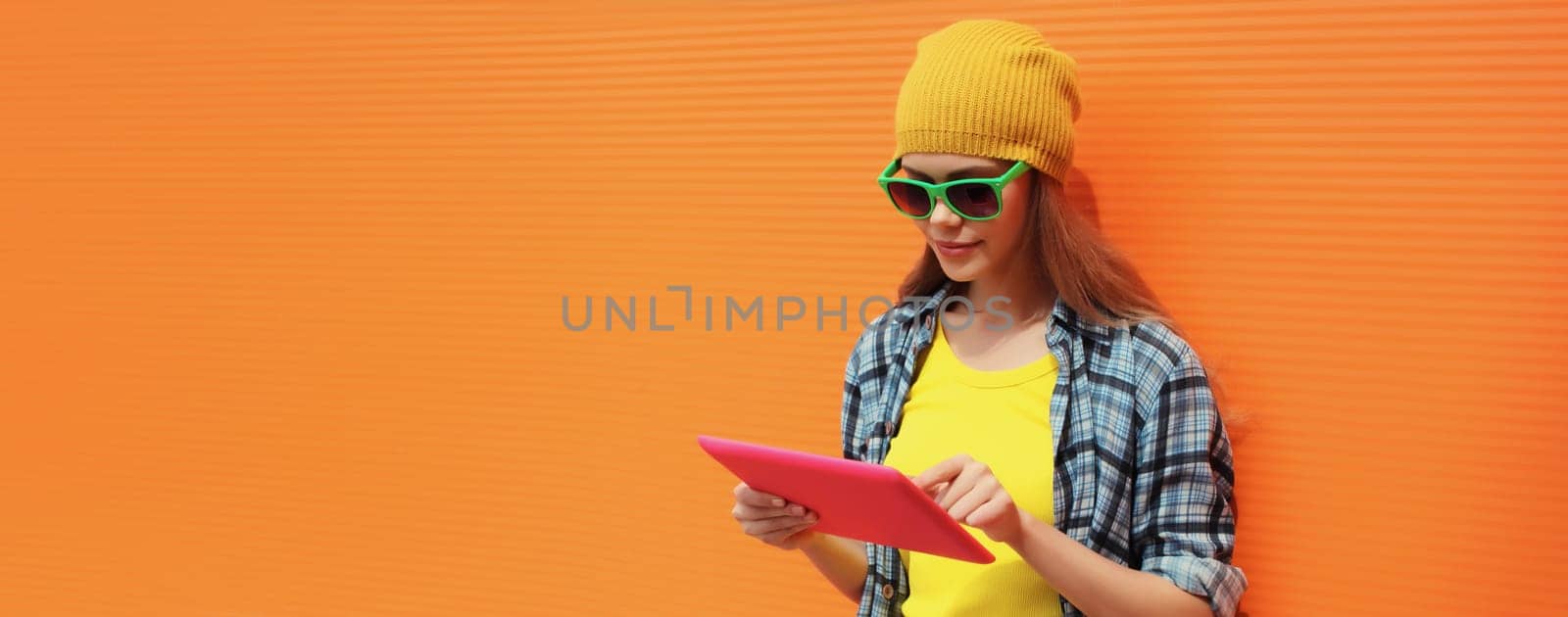 Modern stylish young woman using digital tablet computer looking at device in colorful clothes on city street on orange wall background, blank copy space