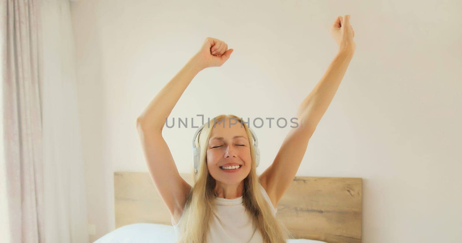 Happy cheerful middle-aged woman listening to music with headphones having fun on the bed at home