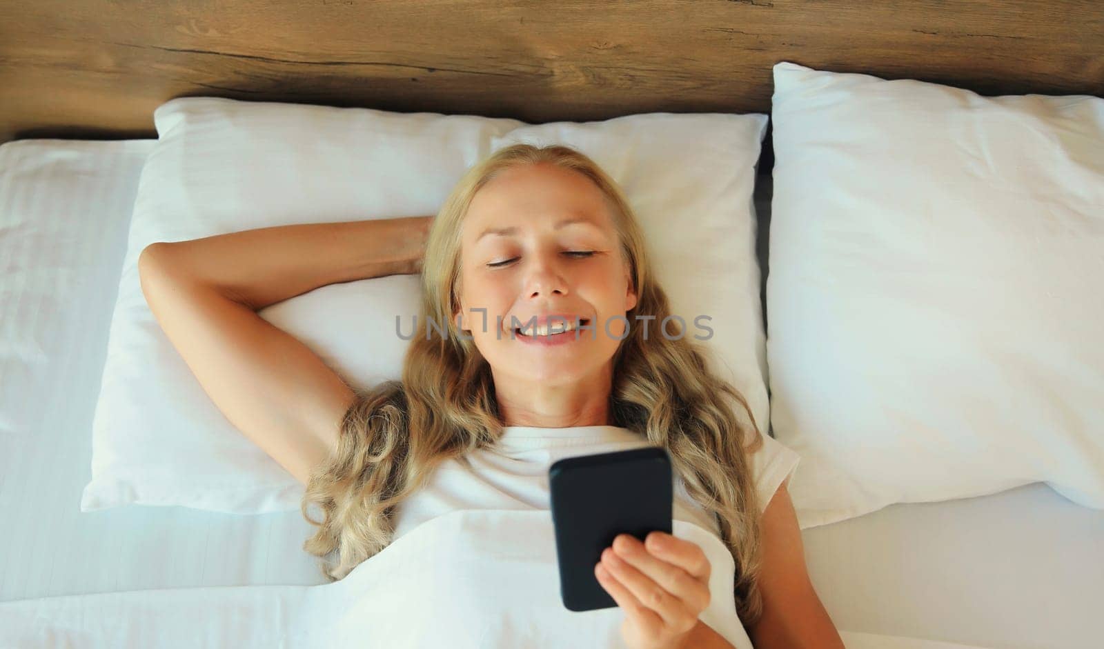 Happy middle aged woman with mobile phone lying on the bed in bedroom at home by Rohappy