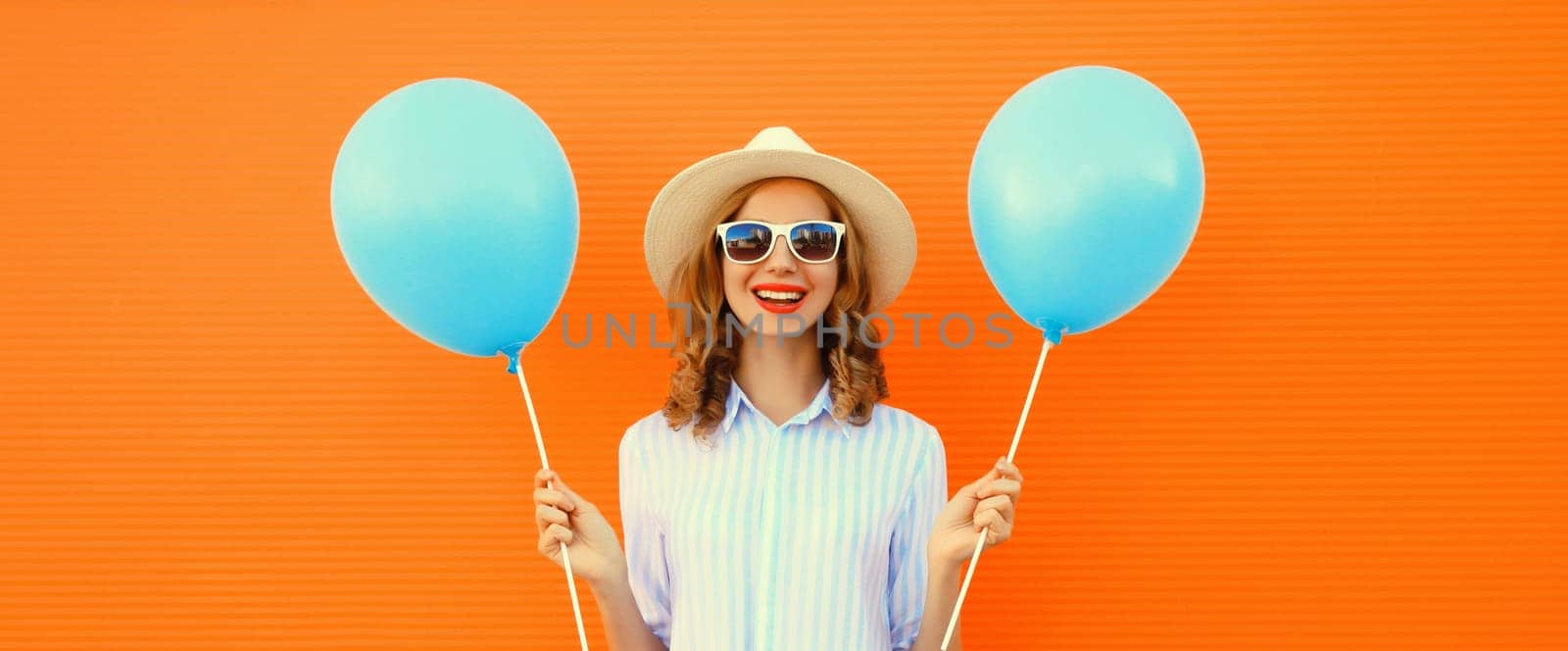 Portrait of happy smiling young woman with blue balloon wearing summer straw hat on orange background