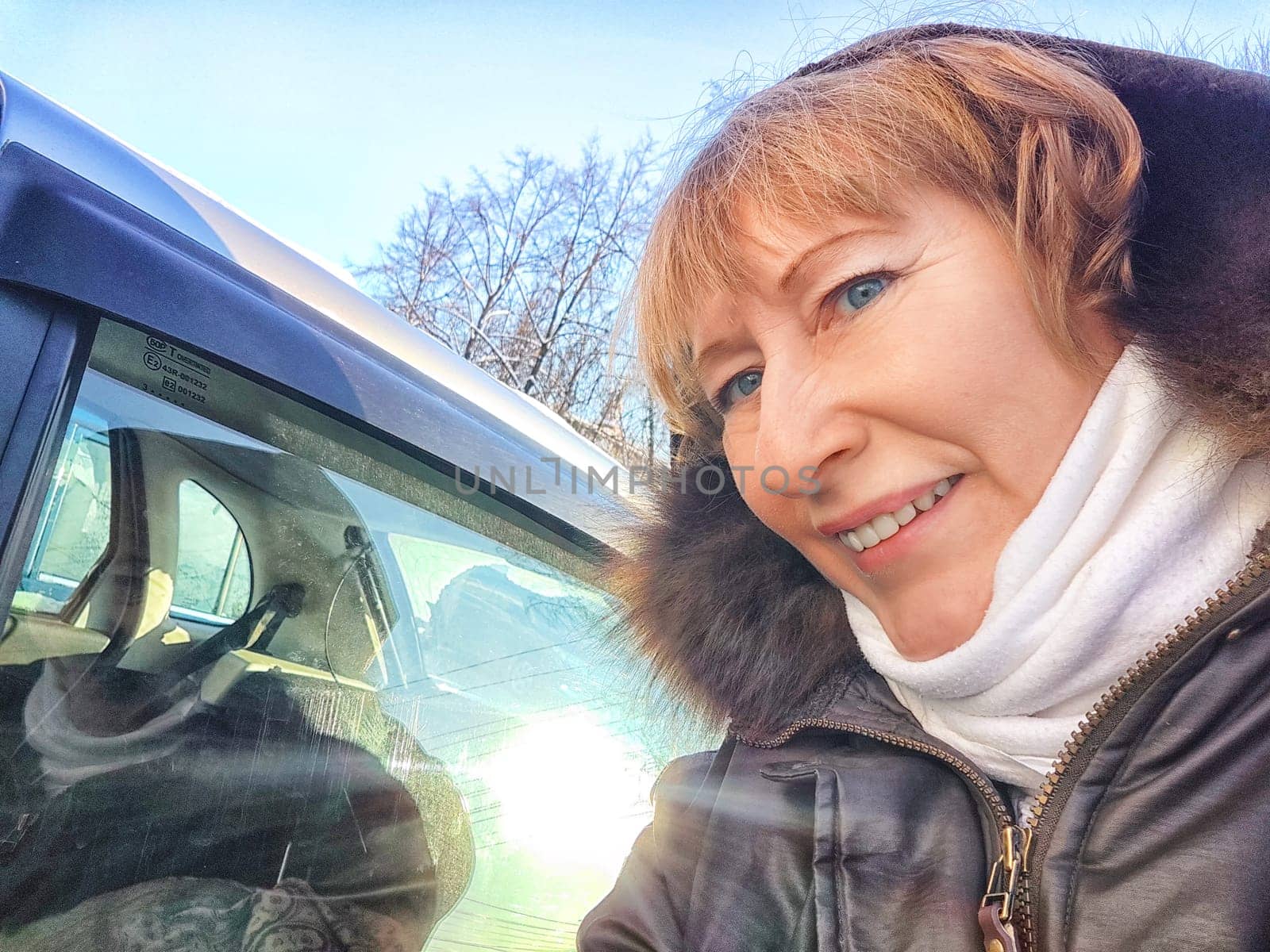 A woman with short hair is smiling at the camera while standing next to a car under clear blue skies, with the sun shining brightly