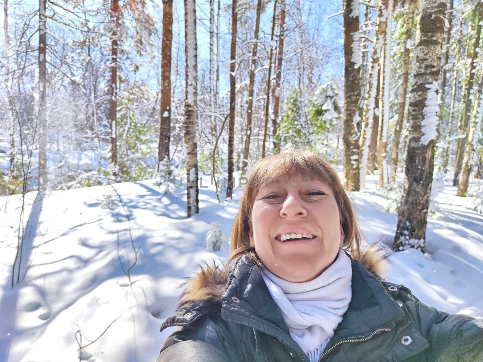 A cheerful middle aged woman in a winter coat taking selfie on nature outdoors in sunny day with blue sky