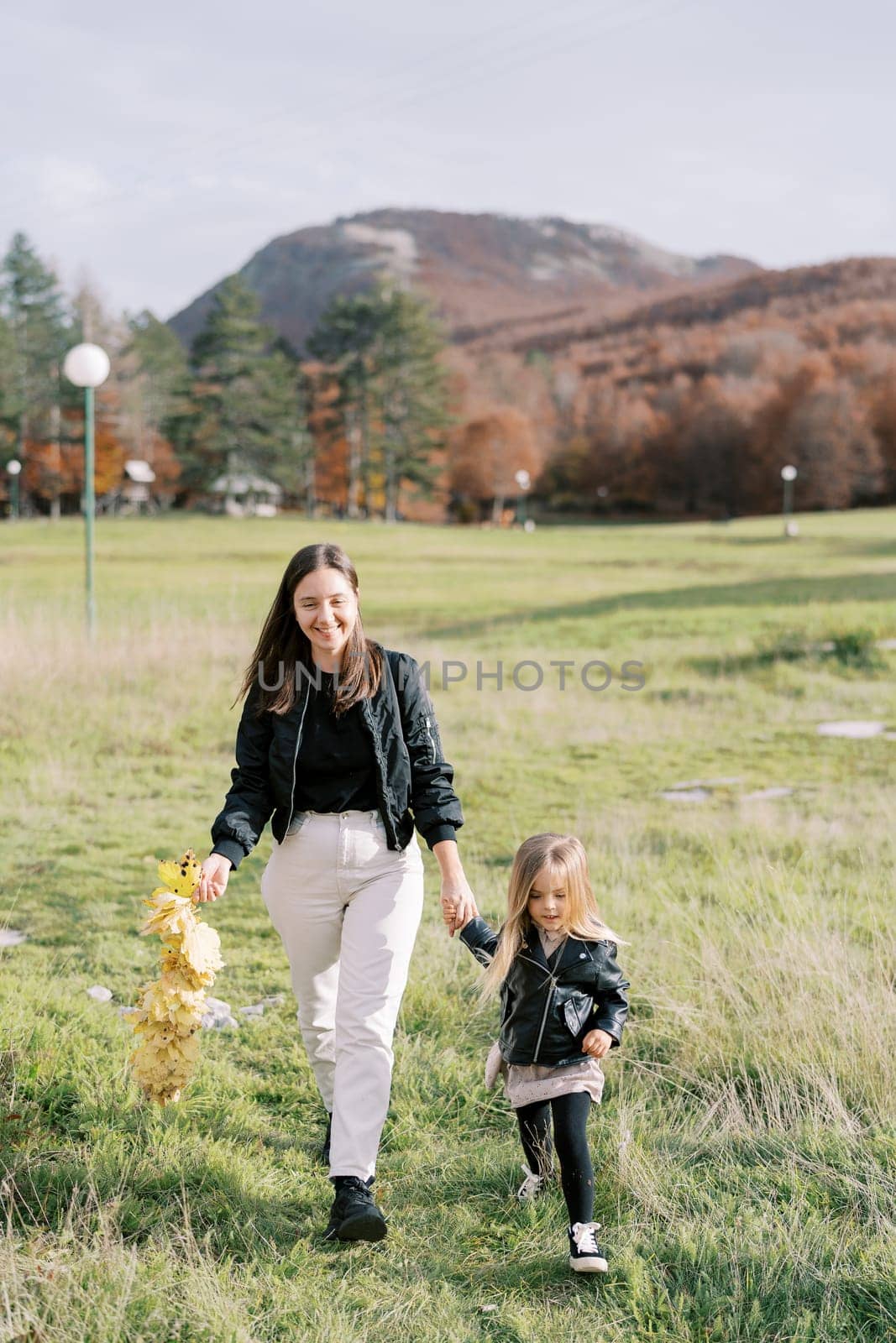 Smiling mother with a wreath of yellow leaves in her hand walks with little girl across the lawn holding hands by Nadtochiy