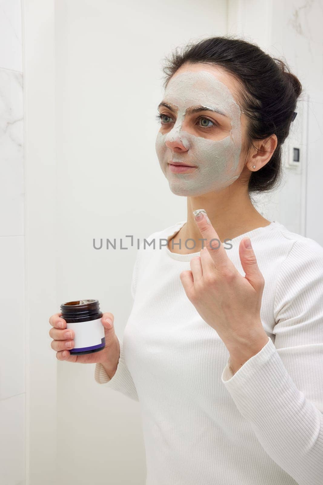 Caucasian woman looking in mirror and applying face mask white in bathroom