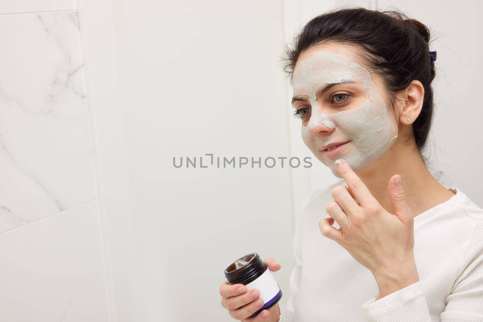 Caucasian woman looking in mirror and applying face mask white in bathroom. copy space