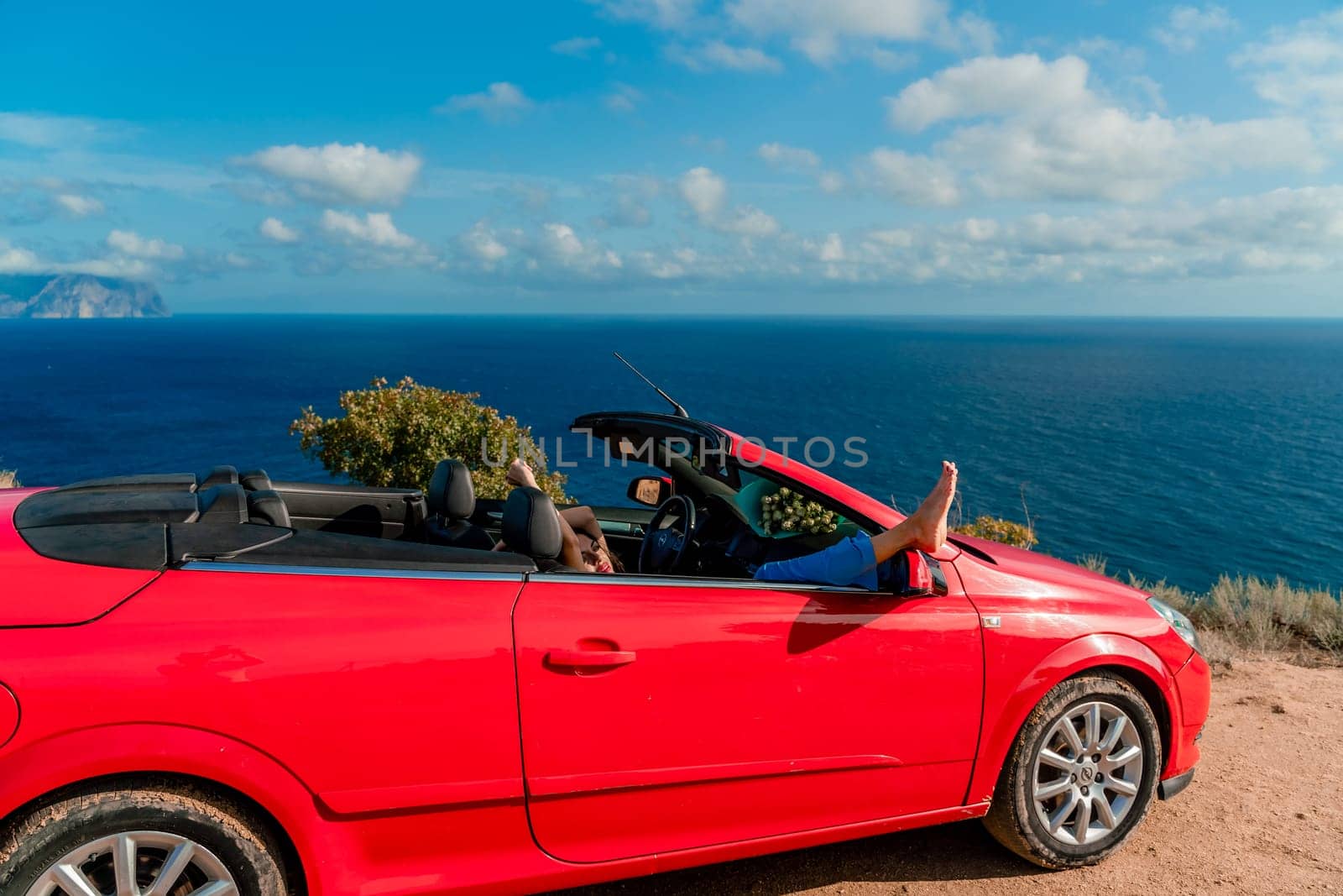 A woman is laying on her feet in a red car, looking out the window at the ocean.