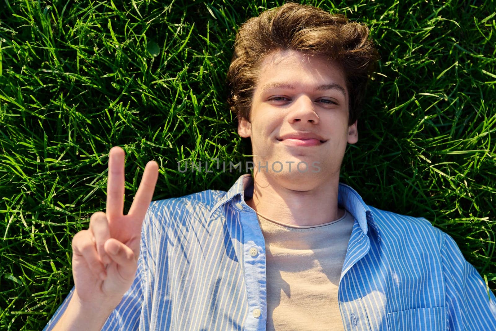 Top view, happy handsome smiling young male looking at camera, lying on background of green lawn grass, showing hand gesture peace victory success