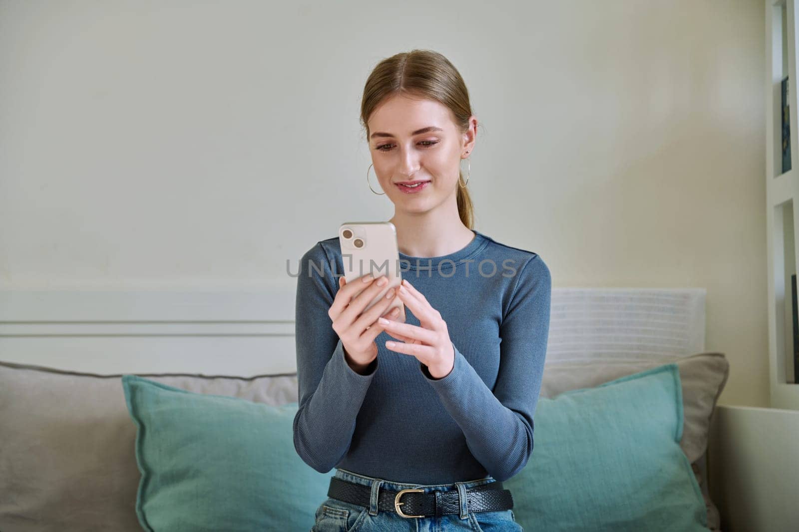 Happy smiling female teenager using smartphone, sitting on couch at home, girl 16,17, 18 years old texting reading messages. Modern digital technologies for communication, leisure, learning, shopping