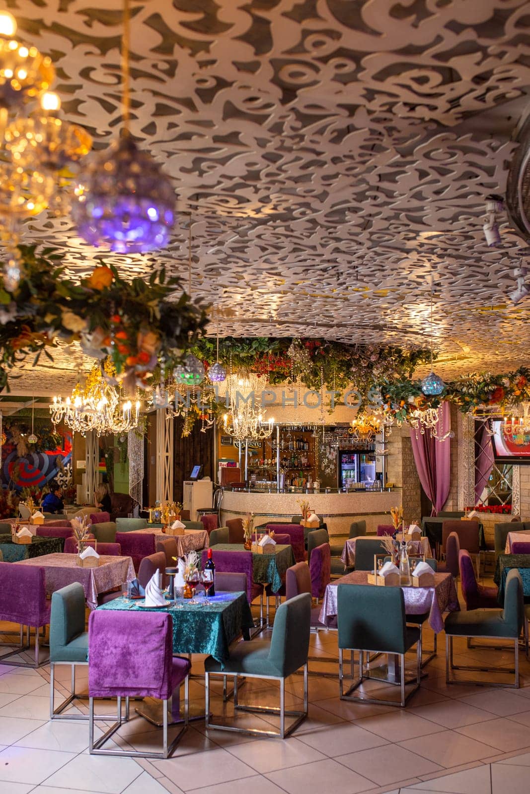 Elegant restaurant interior with round tables, purple tablecloths, chandeliers, and floral decor. Wine bottle on the table.