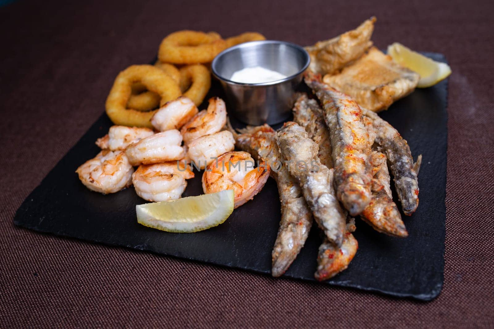 A delicious and healthy meal of fried seafood, including shrimp, fish, and calamari, served with lemon wedges on a black slate plate.