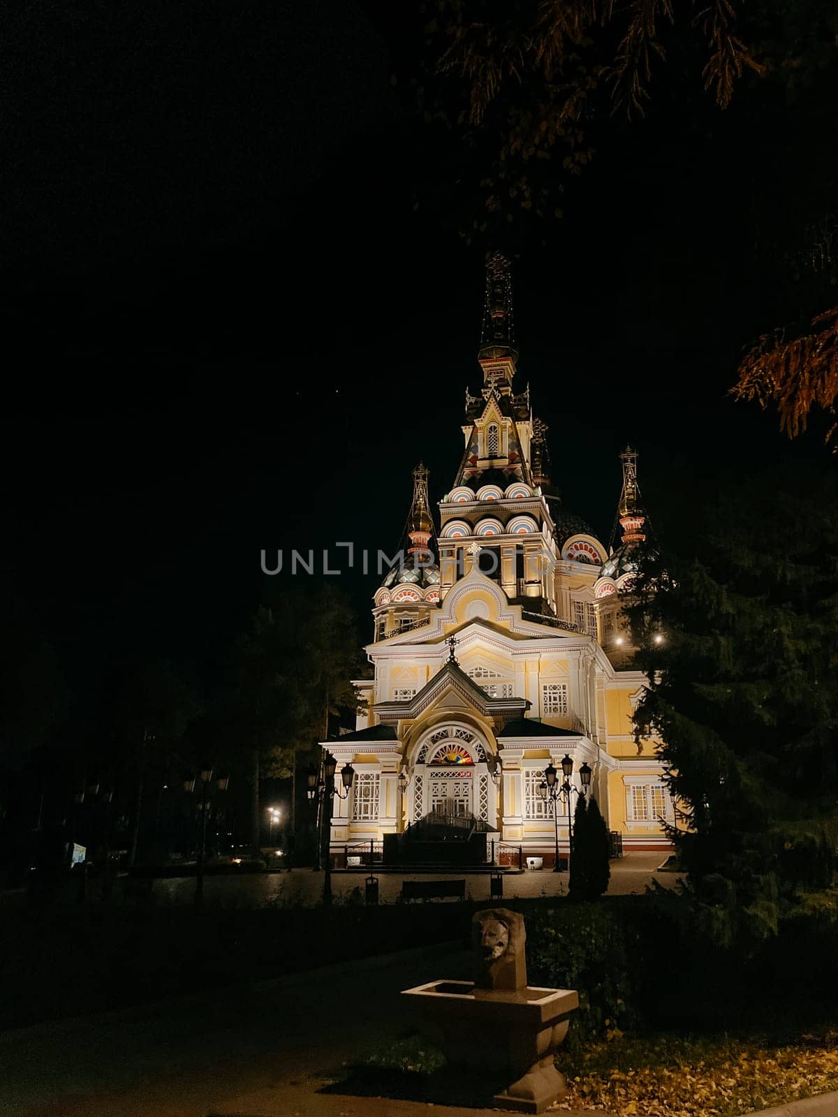 A stunning Russian Orthodox church illuminated at night, with a vibrant yellow exterior, towering steeple, surrounded by trees and park.