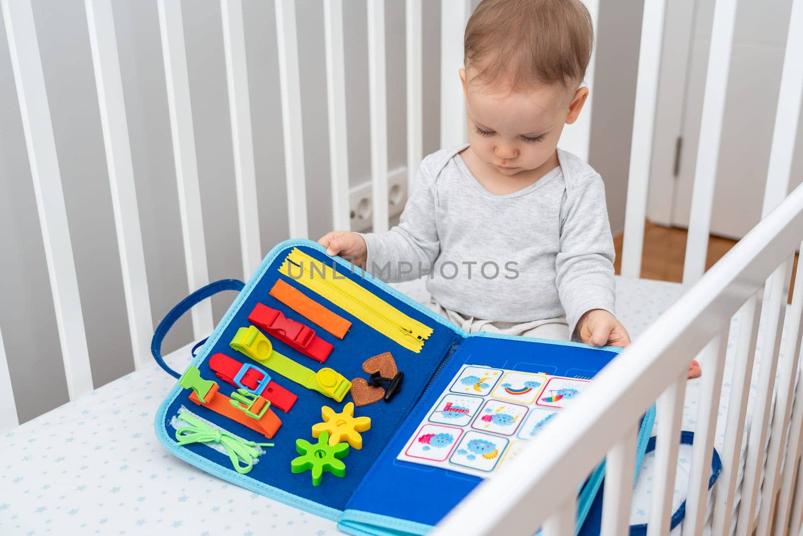 Little baby girl playing with busy book while sitting in crib. Concept of quiet books and modern educational toys by Mariakray