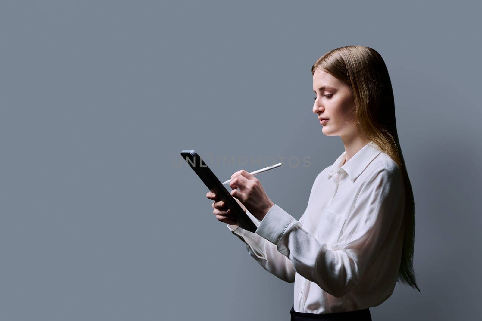Teenage female student using digital tablet on gray background, profile view. Teenager girl 16,17, 18 years old writing with stylus. Education Internet technology e-learning educational services