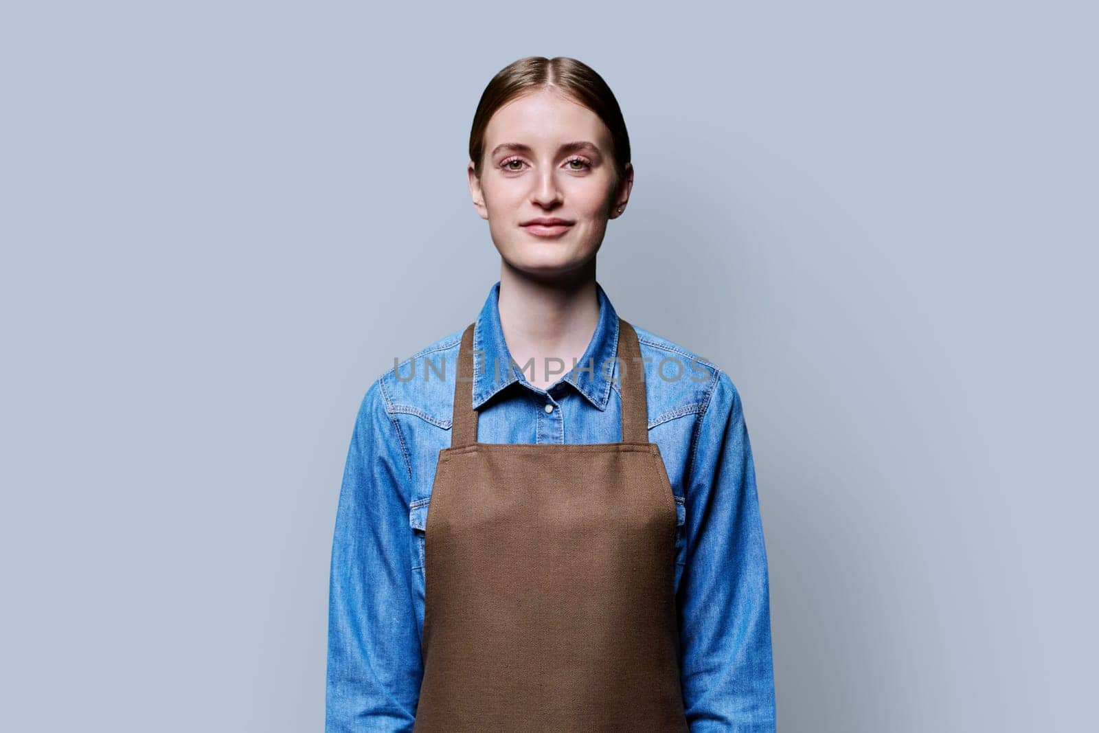 Portrait of young smiling confident woman in apron on grey studio background. Successful positive female looking at camera. Worker, startup, small business, service sector, staff, youth concept