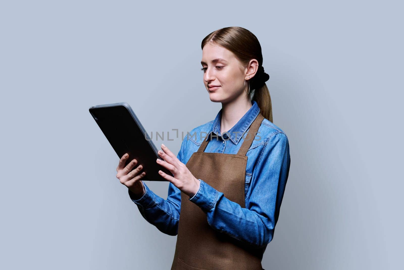 Young female worker in apron holding digital tablet on gray studio background by VH-studio
