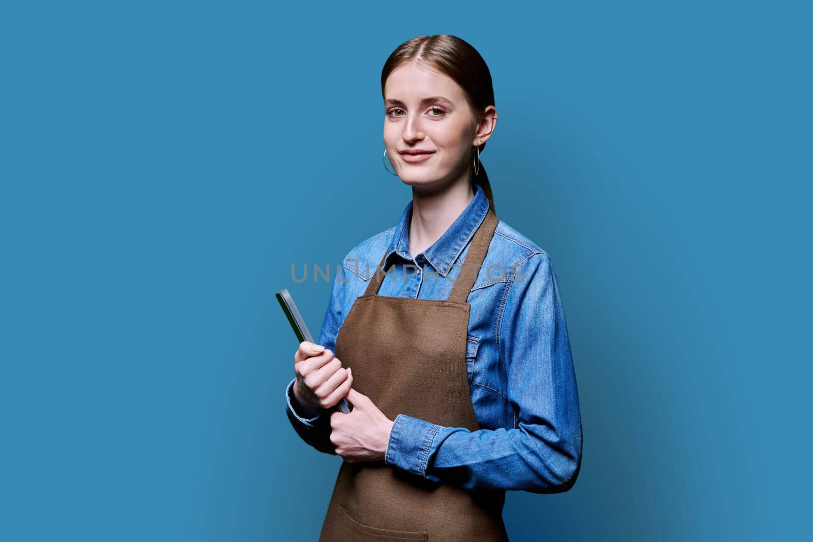 Young smiling female worker in apron uniform holding digital tablet on blue studio background. Digital technologies in business work, Internet apps applications for online orders and customer service