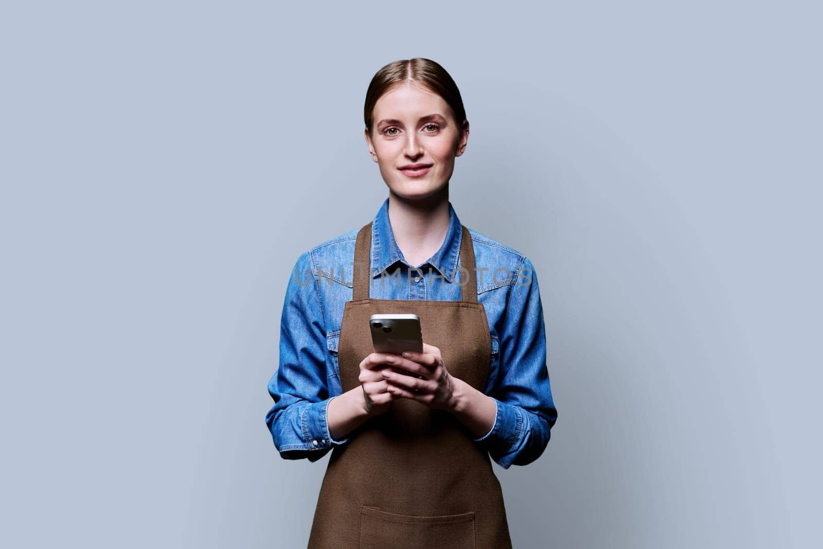 Young female worker in an apron using smartphone on gray studio background by VH-studio